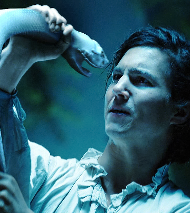 Nächtliche Stimmung. Der Wald im Hintergrund ist nur zu ahnen. Endres, gespielt von Tim Oliver Schultz, im weiten, hellen Hemd, hat die weiße Schlange gepackt und fixiert angespannt ihren Kopf, der sich nur wenige Zentimeter vor seinem Gesicht befindet. Die beiden reden miteinander.