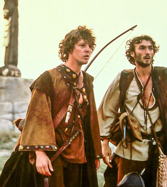 Prinz Martin (Remi Martin) steht zusammen mit seinem Diener Johannes (Valdimir Hajdu) auf einer Wiese und blickt in die Ferne. Der Prinz trägt einen Bogen auf dem Rücken und eine gefüllte Tasche. Beide sind zur Jagd ausgerüstet.