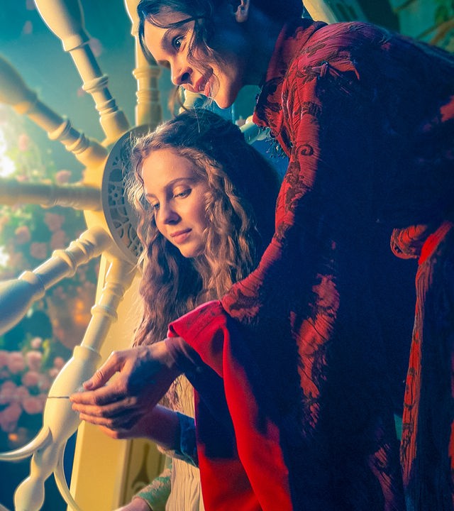 Die Prinzessin Rosabella sitzt mit der bösen Fee Rubia vor einem Spinnrad, Rubia führt der Prinzessin die Hand. Im Hintergrund scheint Licht durch eine Fensterrosette.