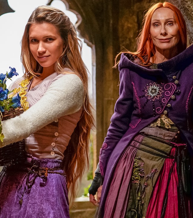 In einem alten, steinernen Gemäuer stehen Rapunzel, gespielt von Anne-Lena Schwing, und Zauberin Eleonor, gespielt von Andrea Sawatzi, vor einem steinernen Fensterrahmen und schauen in die Kamera.