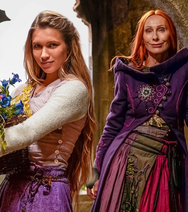 In einem alten, steinernen Gemäuer stehen Rapunzel, gespielt von Anne-Lena Schwing, und Zauberin Eleonor, gespielt von Andrea Sawatzi, vor einem steinernen Fensterrahmen und schauen in die Kamera.