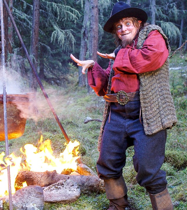 Rumpelstilzchen, verkörpert von Katharina Thalbach, tanzt mit hämischem Blick im Wald um einen dampfenden Kessel herum, der über einer Feuerstelle hängt.