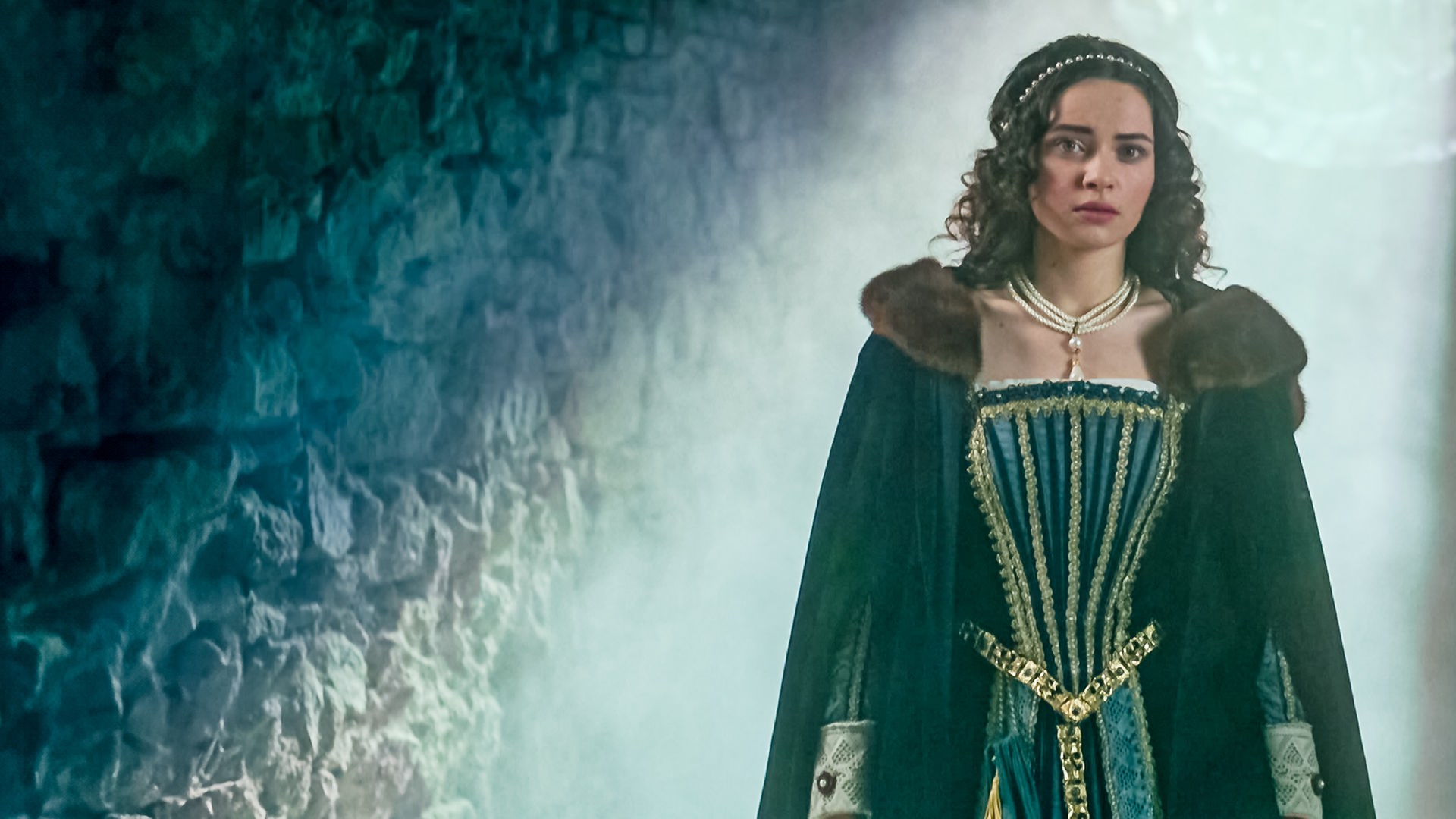 Julia (Katarína Krajčovičová) steht in einem steinernen Tunnel. Skeptisch schaut sie nach vorne. Sie trägt einen dunkelblauen Umhang mit braunem Pelzkragen und ein reich verziertes Kleid. In ihren dunklen langen Haaren sind Perlen eingearbeitet.