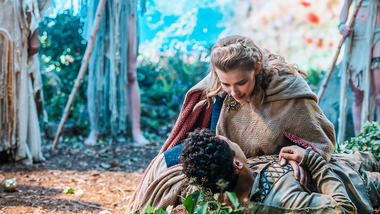 Eine Frau in mittelalterlichen Gewändern, gespielt von Milena Tscharntke, kniet am Boden und hält einen Mann, gespielt von Jan Liem, in ihren Armen. Beide schauen sich an. Im Hintergrund sind im Halbkreis drei Beinpaare von feenhaft gekleideten Personen zu sehen.