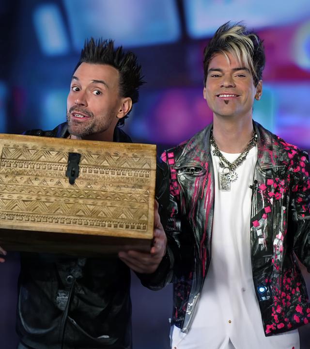 Die Ehrlich Brothers sitzen nebeneinander. Andreas hält eine Holzkiste in die Kamera. Chris lacht dabei in die Kamera.