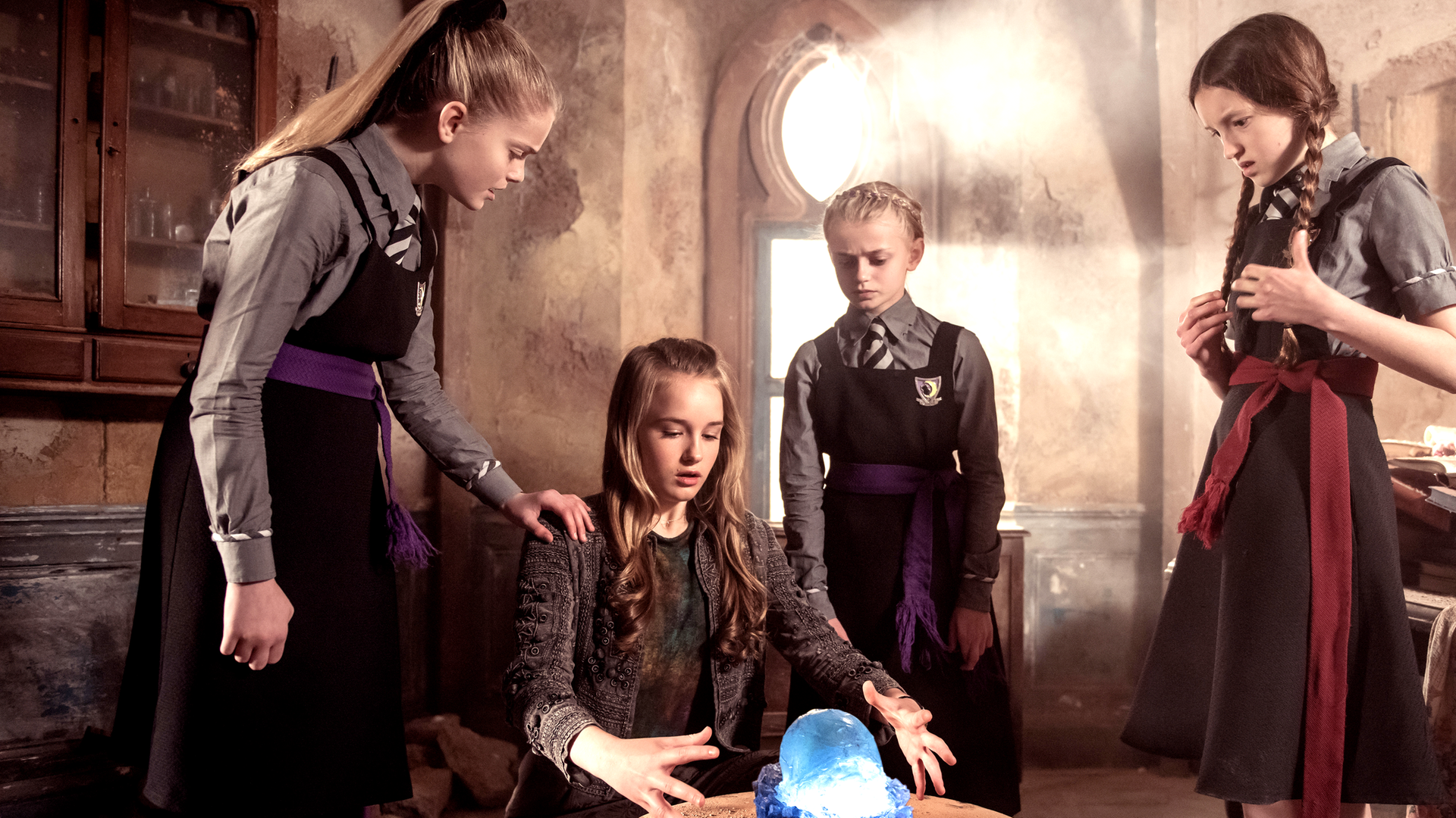 Der Grundstein hat seine Kraft verloren und schimmert blau. Die Hexen Mildred, Esther und zwei Freundinnen stehen um den Stein herum und hantieren mit ihren Händen.