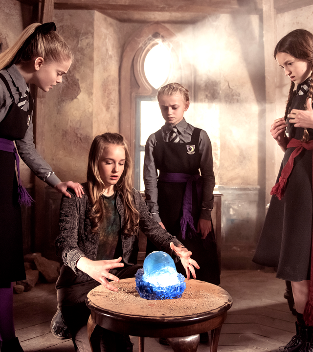 Der Grundstein hat seine Kraft verloren und schimmert blau. Die Hexen Mildred, Esther und zwei Freundinnen stehen um den Stein herum und hantieren mit ihren Händen.