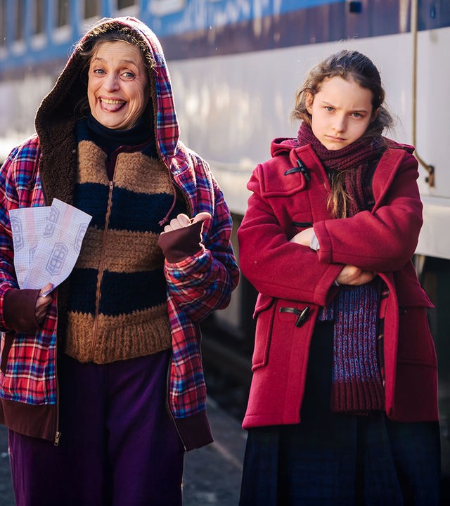Vor einem blau-weißen Waggon präsentiert Engelsanwärterin Addi (Katharina Thalbach) siegesgewiss zwei Zugfahrkarten, die sie für die Reise mit ihrem Schützling benötigt. Marie (Chloé Heinrichs), rechts neben ihr, hat missmutig die Arme verschränkt.