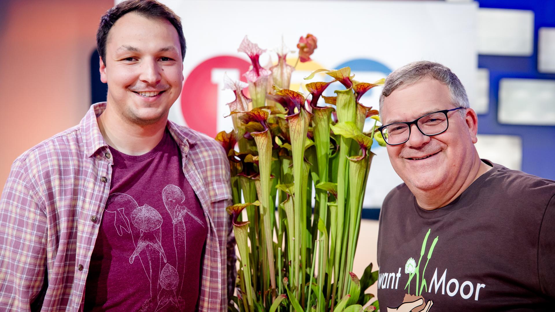 "1, 2 oder 3 - Magische Moore": Tim Biking und Elton stehen im Studio vor dem "1, 2 oder 3"- Logo. Sie halten eine fleischfressende Pflanze zwischen sich.