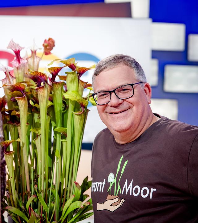 "1, 2 oder 3 - Magische Moore": Tim Biking und Elton stehen im Studio vor dem "1, 2 oder 3"- Logo. Sie halten eine fleischfressende Pflanze zwischen sich.