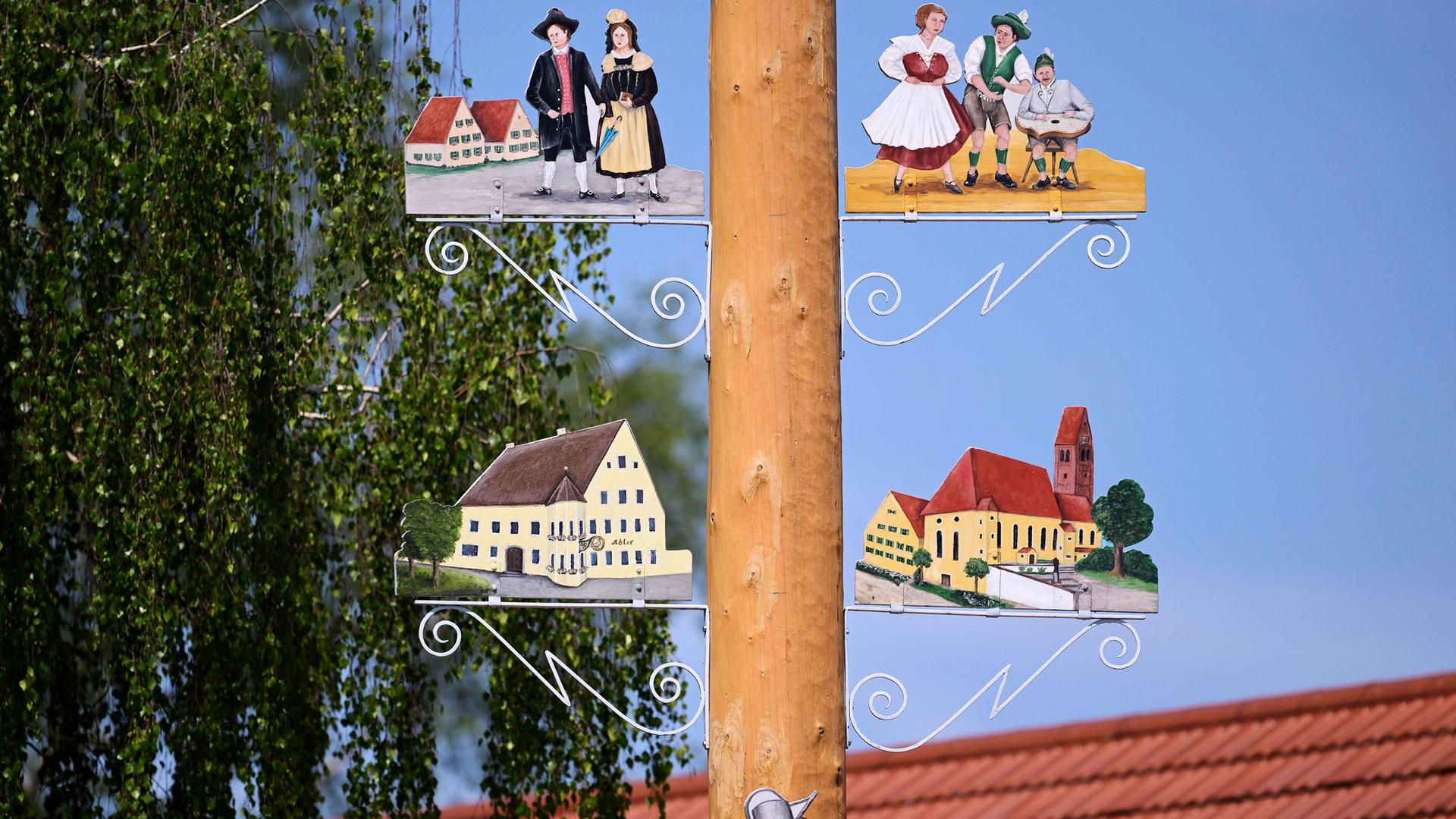 Detail eines Maibaums. Rechts und links vom Stamm sind bemalte Schilder mit Personen in historischen Kleidern und Gebäude aus dem Ort zu sehen.