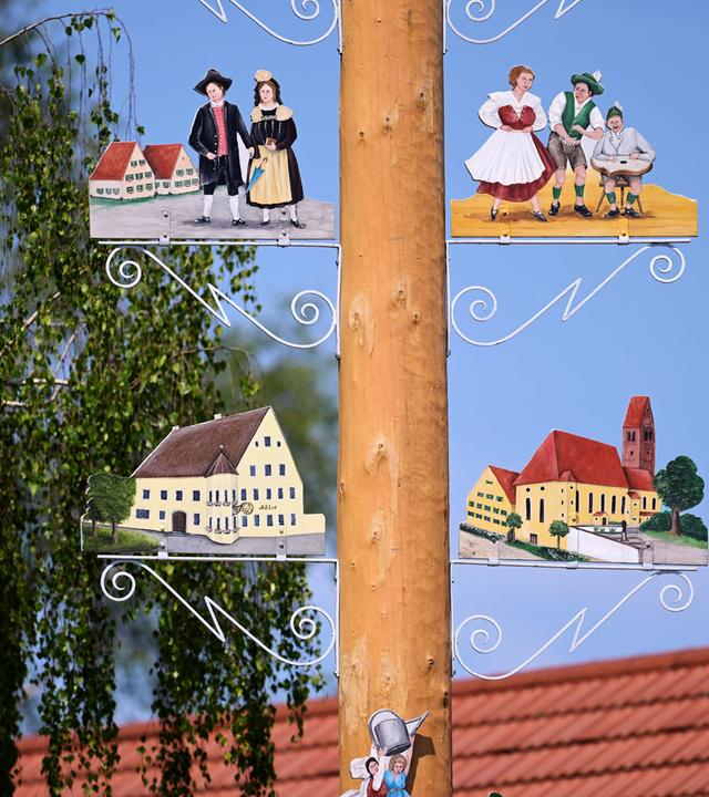 Detail eines Maibaums. Rechts und links vom Stamm sind bemalte Schilder mit Personen in historischen Kleidern und Gebäude aus dem Ort zu sehen.