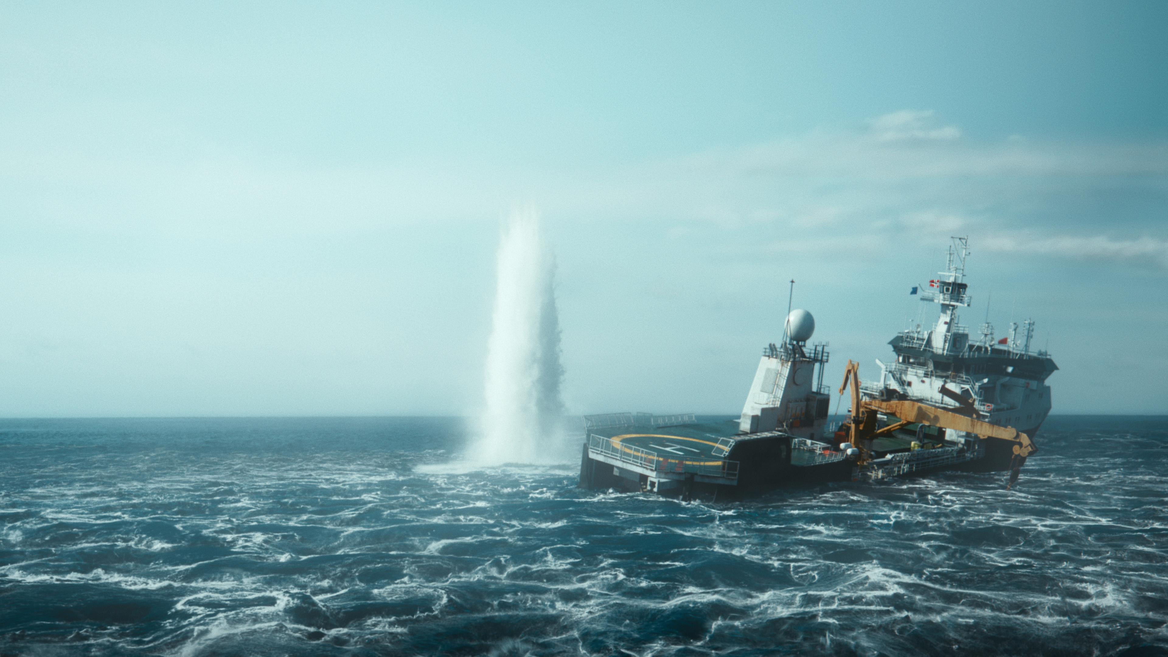 Ein Schiff auf dem Meer. Links davon Fontäne wie von einer Unterwasserexplosion. 