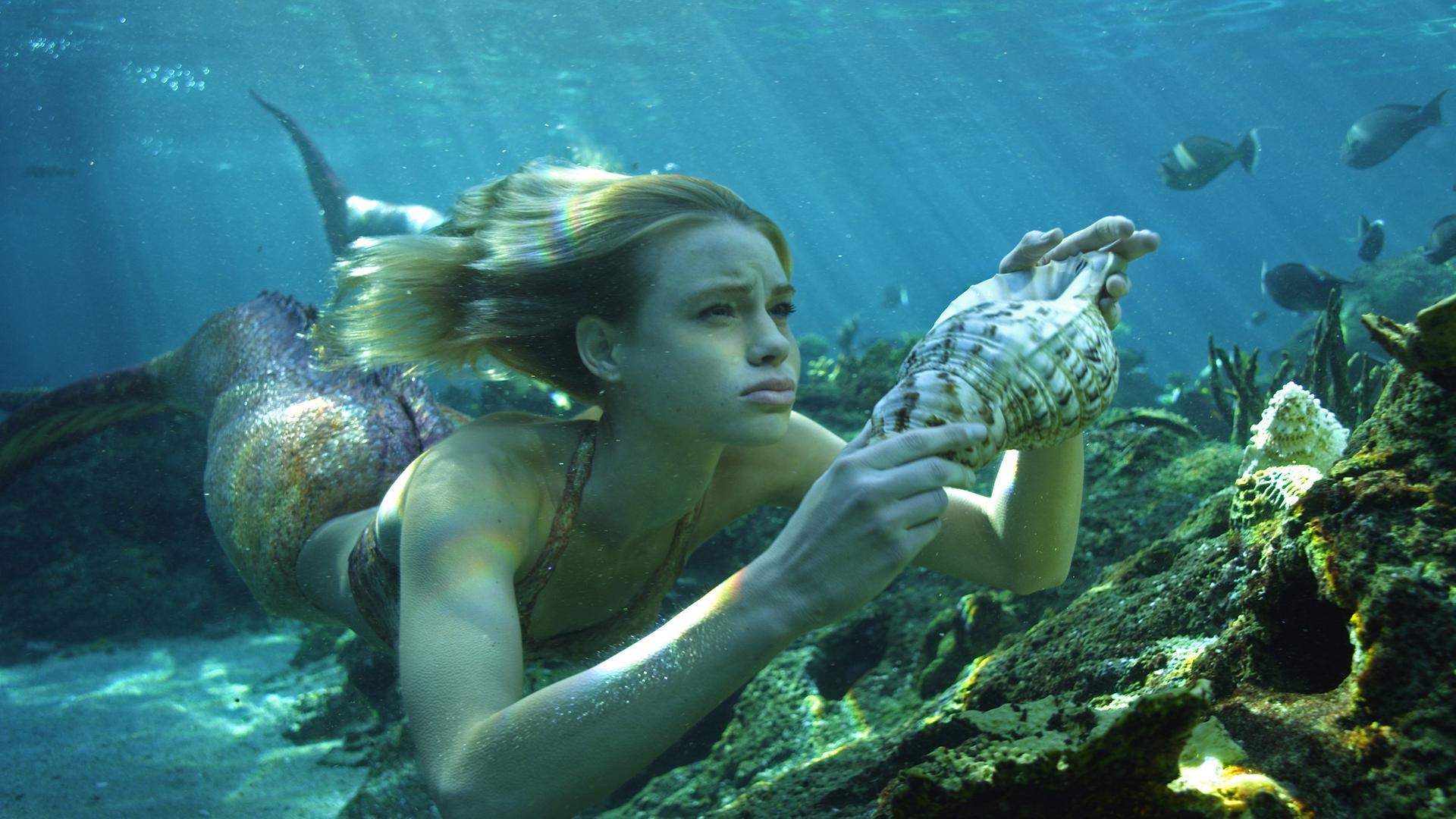 Nixe Lyla schwimmt unter Wasser, dabei hält sie eine Muschel in der Hand. Um sie herum schwimmen Fische.