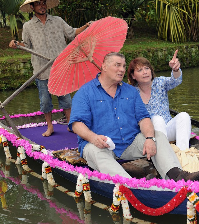 Paul (Heinz Hoenig) und Maria (Marie Gruber) sitzen in einem mit Blumengirlanden geschmückten Ruderboot. Marie hält einen Sonnenschirm und zeigt mit dem Finger auf etwas in der Ferne. Paul blickt dorthin. Hinter ihnen steht ein einheimischer Bootsführer am Ruder.