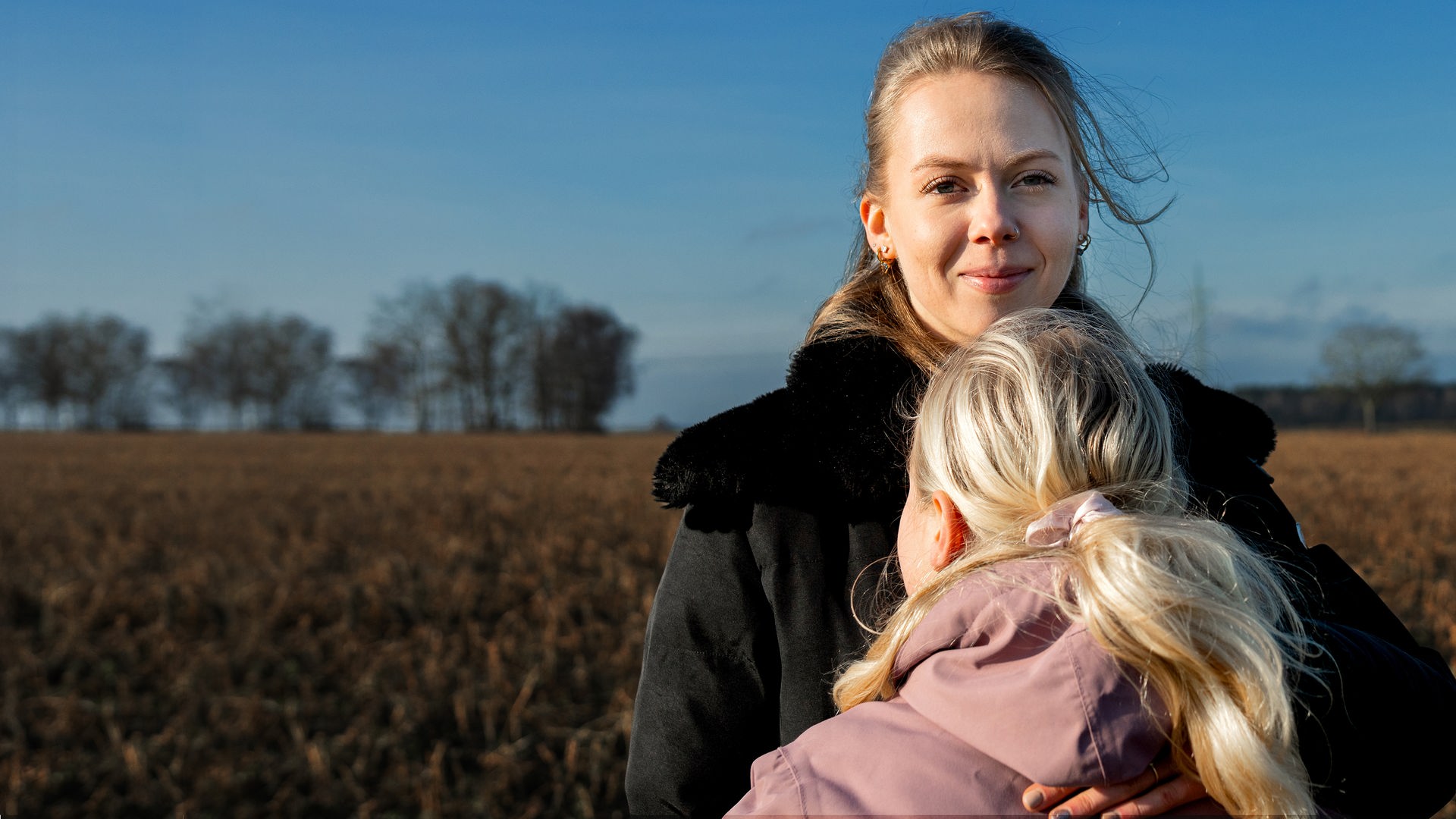 Merle hält ihre Schwester im Arm, die man nur von hinten sieht. Merle schaut lächelnd in die Kamera