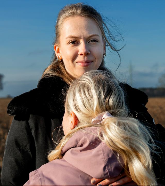 Merle hält ihre Schwester im Arm, die man nur von hinten sieht. Merle schaut lächelnd in die Kamera