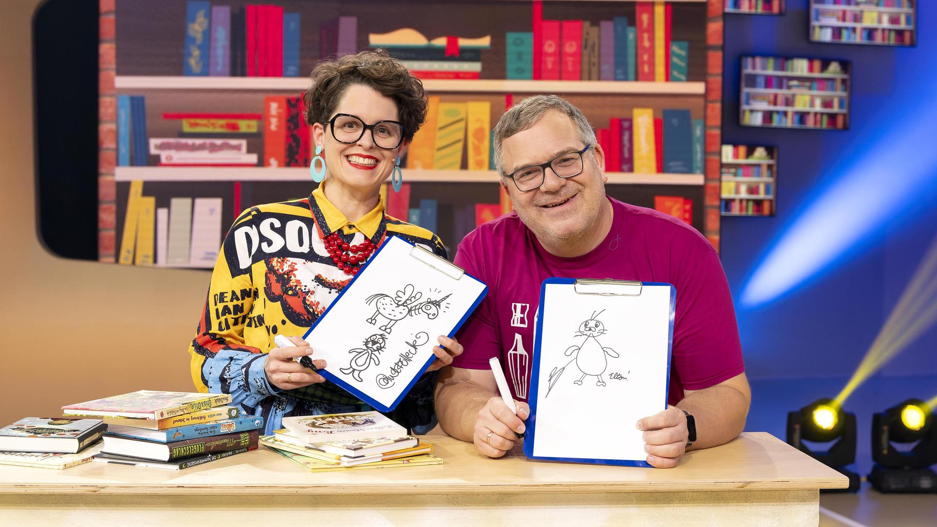 Illustratorin Nina Dulleck und Elton sitzen lächelnd hinter einem Tisch, auf dem mehrere Bücher liegen. Beide halten Zeichnungen in den Händen. Im Hintergrund steht ein volles Bücherregal.