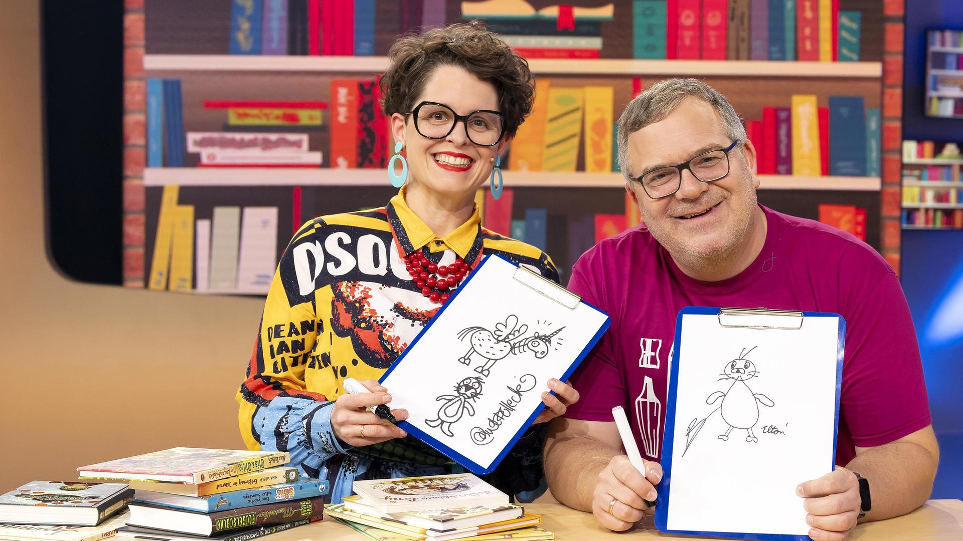 Illustratorin Nina Dulleck und Elton sitzen lächelnd hinter einem Tisch, auf dem mehrere Bücher liegen. Beide halten Zeichnungen in den Händen. Im Hintergrund steht ein volles Bücherregal.