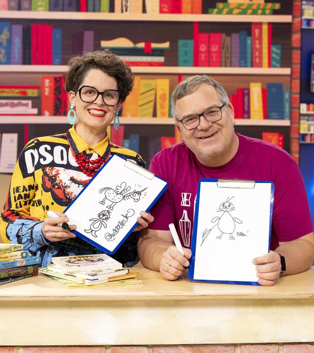 Illustratorin Nina Dulleck und Elton sitzen lächelnd hinter einem Tisch, auf dem mehrere Bücher liegen. Beide halten Zeichnungen in den Händen. Im Hintergrund steht ein volles Bücherregal.