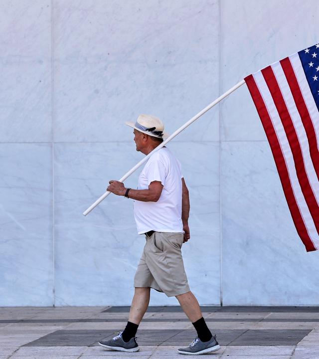 Ein Mann läuft mit einer US-Flagge auf der Schulter vor einer grauen Steinwand.