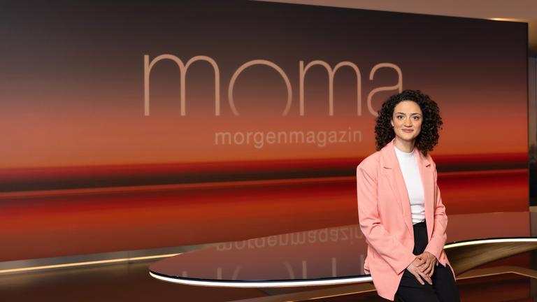 Maral Bazargani, neue Moderatorin des ZDF-Morgenmagazins, lehnt im Studio vor einer großen, warm beleuchteten Hintergrundwand mit der Aufschrift „moma“ an einem Tisch. Sie trägt einen hellen Blazer über einem weißen Oberteil und blickt mit einem leichten Lächeln in die Kamera