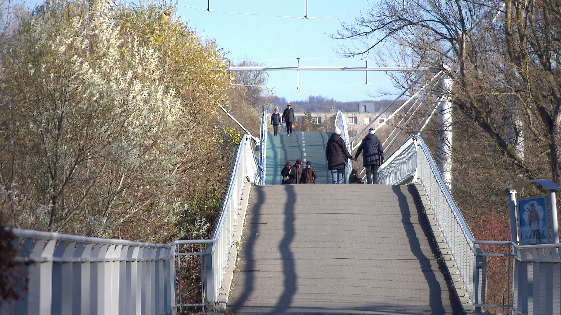 Das Bild zeigt eine Fahrrad- und Fußgängerbrücke, die von Marchegg in die Slowakei führt. Die Brücke hat eine gebogene Form und ist aus Metall mit einem glatten Belag. Auf der Brücke sind mehrere Fußgänger zu sehen, die in beide Richtungen gehen. Einige Personen tragen warme Kleidung, da es anscheinend kühl ist.   Auf beiden Seiten der Brücke wachsen Bäume und Sträucher, die teilweise verblühte Blätter zeigen. Im Hintergrund sind einige Gebäude sichtbar, die leicht unscharf sind und aufgrund der Entfernung nicht genau zu erkennen sind. Der Himmel ist klar und blau.   Die gesamte Szene vermittelt eine ruhige Atmosphäre, die den Grenzbereich zwischen Österreich und der Slowakei symbolisiert.