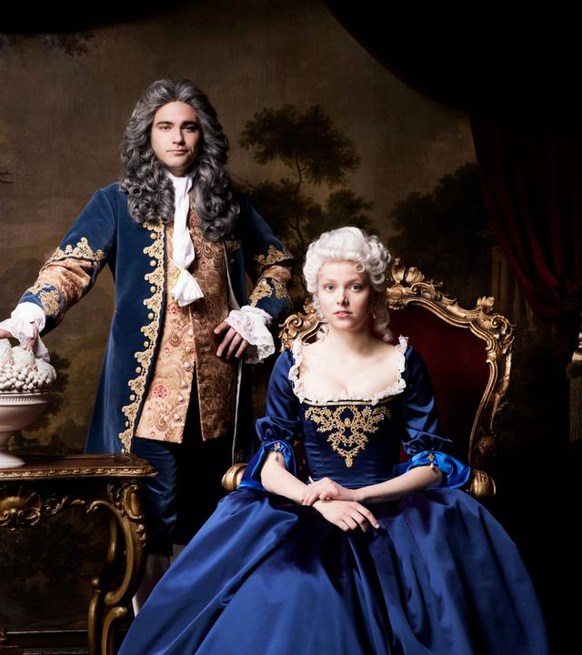 Ein Mann steht neben einer sitzenden Frau in blauer Robe.