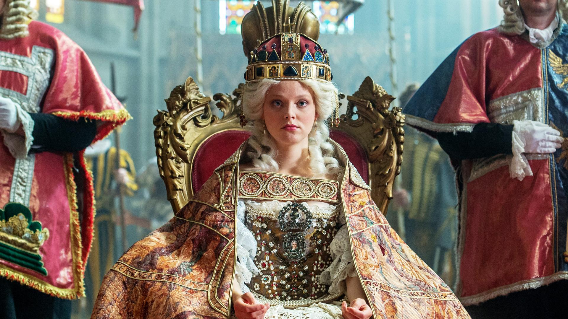 Das Bild zeigt eine Szene aus der Geschichte von Maria Theresia. Im Mittelpunkt sitzt eine junge Frau, die Maria Theresia darstellt, auf einem prunkvollen, goldenen Thron. Sie trägt ein aufwendiges Kostüm mit viel Spitze und Edelsteinen sowie einen prächtigen, auffälligen Hut oder eine Krone auf dem Kopf. Ihr Gesichtsausdruck ist ernst und konzentriert.  Sie ist umgeben von mehreren Personen, die in historischen Gewändern gekleidet sind. Diese Personen stehen zu beiden Seiten des Thrones und scheinen offizielle Würdenträger oder Berater zu sein. Im Hintergrund sind Ansichten von farbigen Fenstern erkennbar, die dem Raum eine feierliche Atmosphäre verleihen.  Die gesamte Szenerie wirkt majestätisch und historisch, was durch die aufwändigen Details der Kleidung und die Gestaltung des Thrones verstärkt wird.