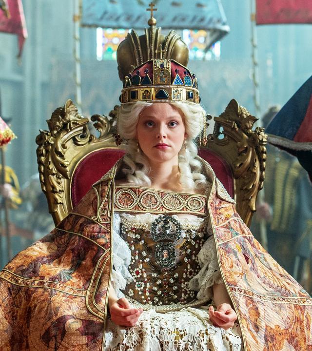 Das Bild zeigt eine Szene aus der Geschichte von Maria Theresia. Im Mittelpunkt sitzt eine junge Frau, die Maria Theresia darstellt, auf einem prunkvollen, goldenen Thron. Sie trägt ein aufwendiges Kostüm mit viel Spitze und Edelsteinen sowie einen prächtigen, auffälligen Hut oder eine Krone auf dem Kopf. Ihr Gesichtsausdruck ist ernst und konzentriert.  Sie ist umgeben von mehreren Personen, die in historischen Gewändern gekleidet sind. Diese Personen stehen zu beiden Seiten des Thrones und scheinen offizielle Würdenträger oder Berater zu sein. Im Hintergrund sind Ansichten von farbigen Fenstern erkennbar, die dem Raum eine feierliche Atmosphäre verleihen.  Die gesamte Szenerie wirkt majestätisch und historisch, was durch die aufwändigen Details der Kleidung und die Gestaltung des Thrones verstärkt wird.