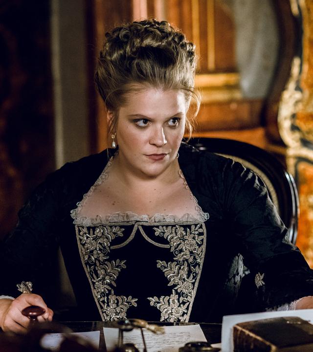 Das Bild zeigt eine Frau in einem historischen Raum, die an einem Tisch sitzt. Sie trägt ein elegantes schwarzes Kleid mit aufwendigen, hellen Stickereien im oberen Bereich. Ihr Haar ist hochgesteckt, und sie hat große, auffällige Ohrringe. Die Mimik der Frau wirkt ernst und nachdenklich, sie scheint auf etwas konzentriert zu sein, während sie auf einem Blatt Papier schaut, das auf dem Tisch vor ihr liegt.   Auf dem Tisch stehen ein Glas Wasser und eine Karaffe, sowie einige Dokumente. Der Hintergrund zeigt Wände mit reich verzierten Holzvertäfelungen und Möbel, die auf eine historische Epoche hinweisen. Die Atmosphäre wirkt majestätisch und zeugt von einem prunkvollen Interieur.