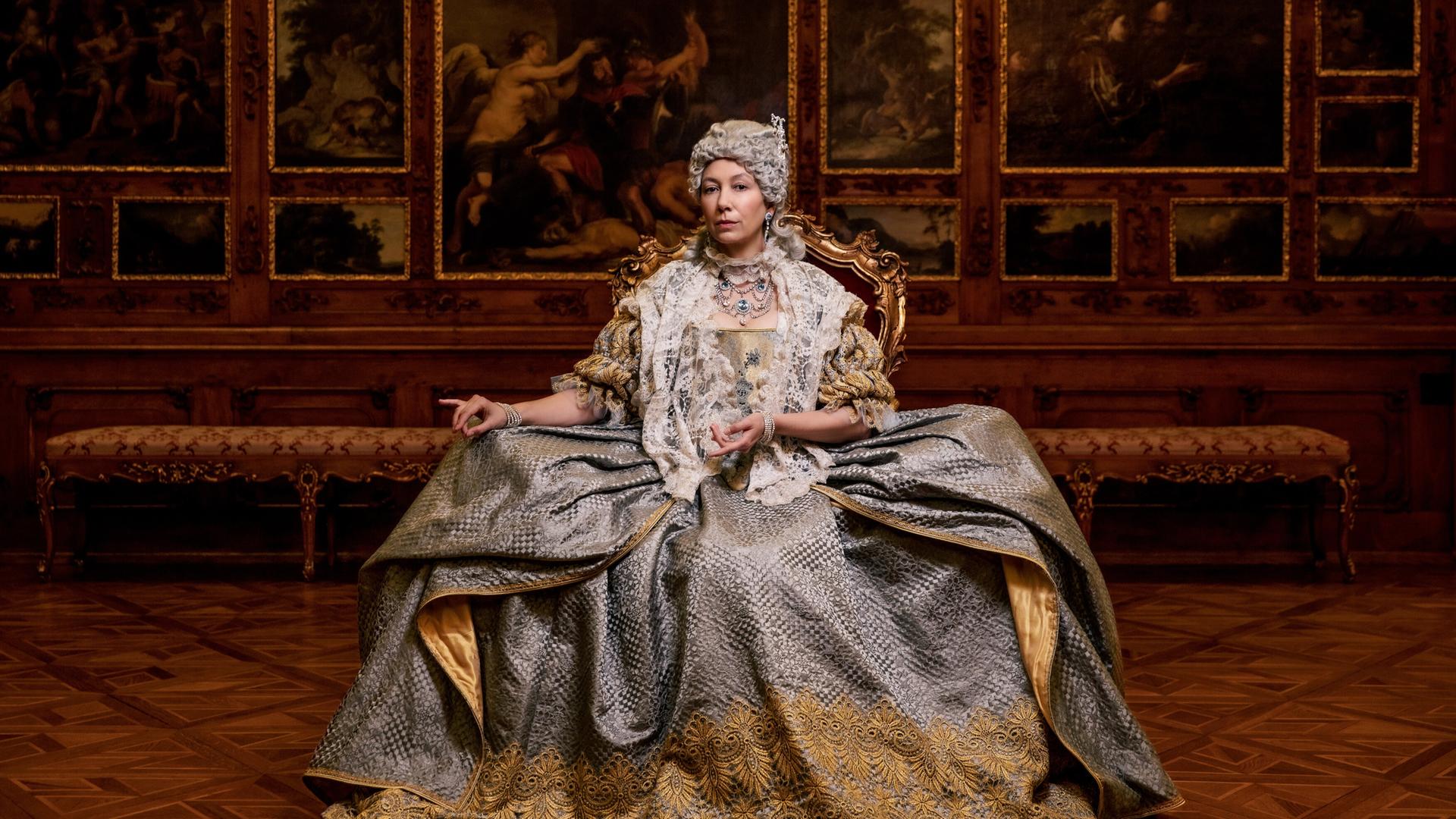Das Bild zeigt eine Frau in historischer Kleidung, die auf einem prächtigen Stuhl sitzt. Sie trägt ein aufwändig gestaltetes Kleid, das in Grautönen gehalten ist und mit goldenen Stickereien verziert ist. Ihr Oberteil ist mit einem weißen Spitzenmantel bedeckt. Die Frau hat eine aufwändige Frisur und Schmuck angelegt, der ihren Hals und ihre Schultern ziert.  Im Hintergrund sind kunstvoll gestaltete Wände mit großen Gemälden, die Szenen aus der Antike oder Mythologie darstellen. Die Wände sind aus dunklem Holz und tragen goldene Verzierungen, die den eleganten, historischen Kontext unterstreichen. Der Boden besteht aus einem auffälligen Holzparkett in einem geometrischen Muster.   Die gesamte Szenerie vermittelt einen Eindruck von Prunk und Macht, passend zu der dargestellten historischen Figur.