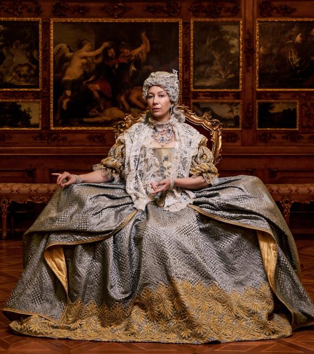 Das Bild zeigt eine Frau in historischer Kleidung, die auf einem prächtigen Stuhl sitzt. Sie trägt ein aufwändig gestaltetes Kleid, das in Grautönen gehalten ist und mit goldenen Stickereien verziert ist. Ihr Oberteil ist mit einem weißen Spitzenmantel bedeckt. Die Frau hat eine aufwändige Frisur und Schmuck angelegt, der ihren Hals und ihre Schultern ziert.  Im Hintergrund sind kunstvoll gestaltete Wände mit großen Gemälden, die Szenen aus der Antike oder Mythologie darstellen. Die Wände sind aus dunklem Holz und tragen goldene Verzierungen, die den eleganten, historischen Kontext unterstreichen. Der Boden besteht aus einem auffälligen Holzparkett in einem geometrischen Muster.   Die gesamte Szenerie vermittelt einen Eindruck von Prunk und Macht, passend zu der dargestellten historischen Figur.