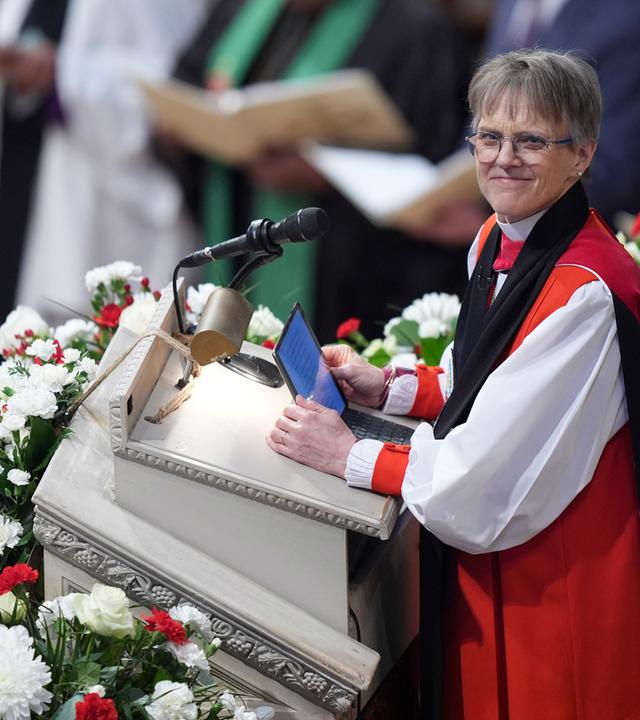 Eine Pfarrerin, gekleidet in eine rote Robe, steht auf einer Blumengeschmückten Kanzel in einer Kirche.