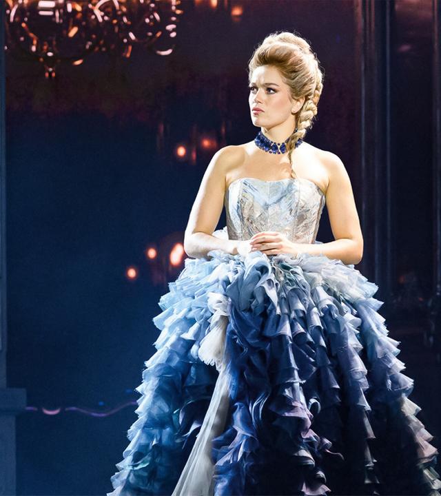 Das Bild zeigt eine Szene aus einem Musical, das sich mit dem Leben von Maria Theresia beschäftigt. Im Vordergrund steht eine weibliche Figur, die eine opulente, mehrschichtige, blau-weiße Robe trägt. Der Unterteil der Robe besteht aus vielen Rüschen, die in verschiedenen Blau- und Grautönen gestaltet sind. Die Figur hat eine geflochtene Frisur und trägt eine auffällige Halskette. Ihr Gesichtsausdruck ist ernst und nachdenklich.   Im Hintergrund sind dunkle Wände zu sehen, die mit eleganten, hochglänzenden Oberflächen und einem dezenten Lichtspiel akzentuiert sind. Einige Lichtquellen scheinen aus dem Hintergrund zu strahlen, was eine dramatische Atmosphäre schafft. Die Szene vermittelt ein Gefühl von Eleganz und Stärke, unterstreicht die historische Thematik des Musicals und reflektiert die Herausforderungen einer starken Frau in einer Machtposition.