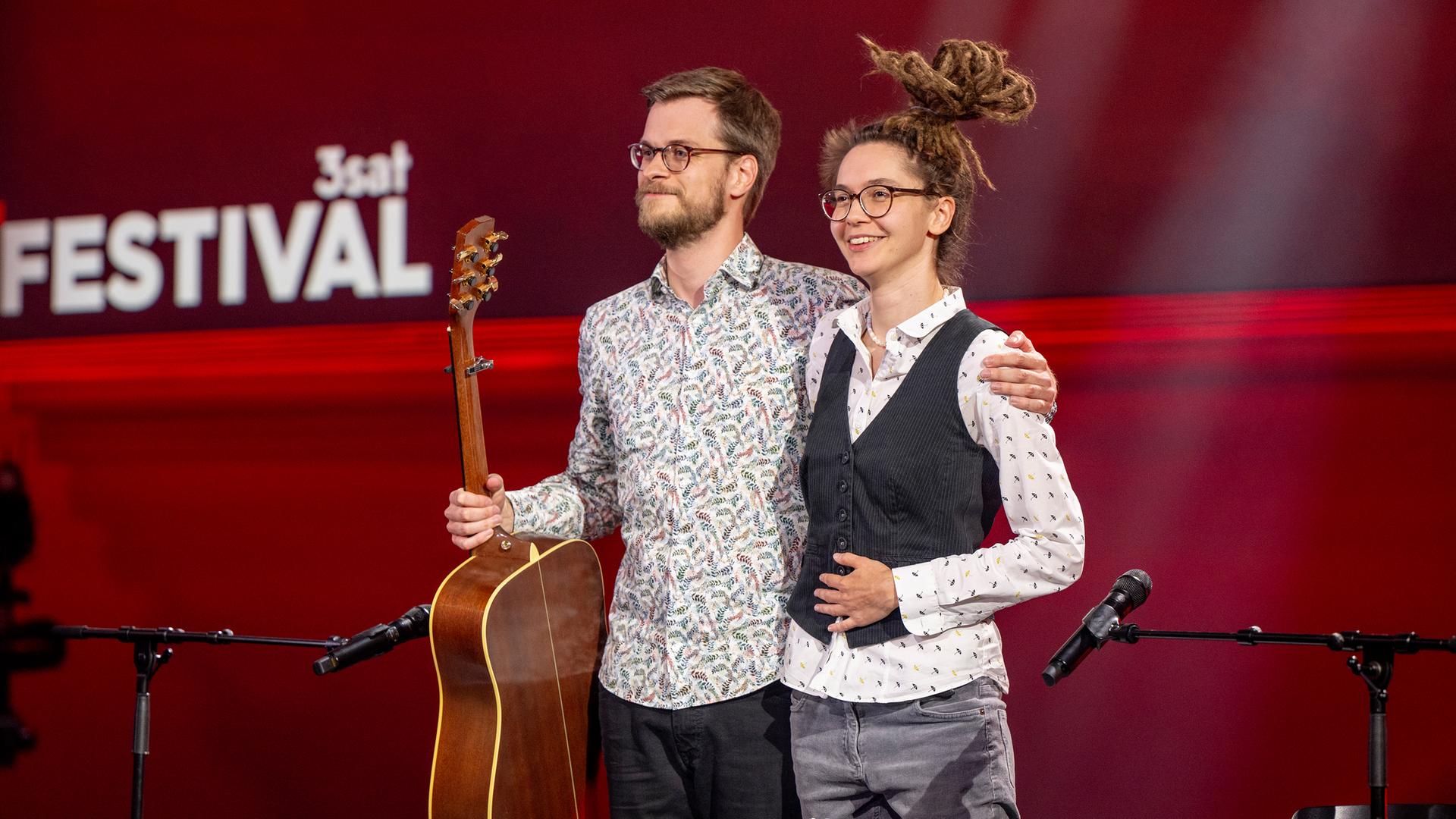 Marie Diot steht auf der rechten Seite des Bildes. Auf der linken Seite ist ihr Gitarrist zu sehen. Der Hintergrund ist rot