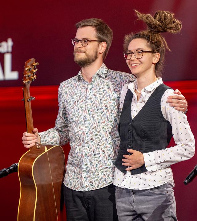 Marie Diot steht auf der rechten Seite des Bildes. Auf der linken Seite ist ihr Gitarrist zu sehen. Der Hintergrund ist rot