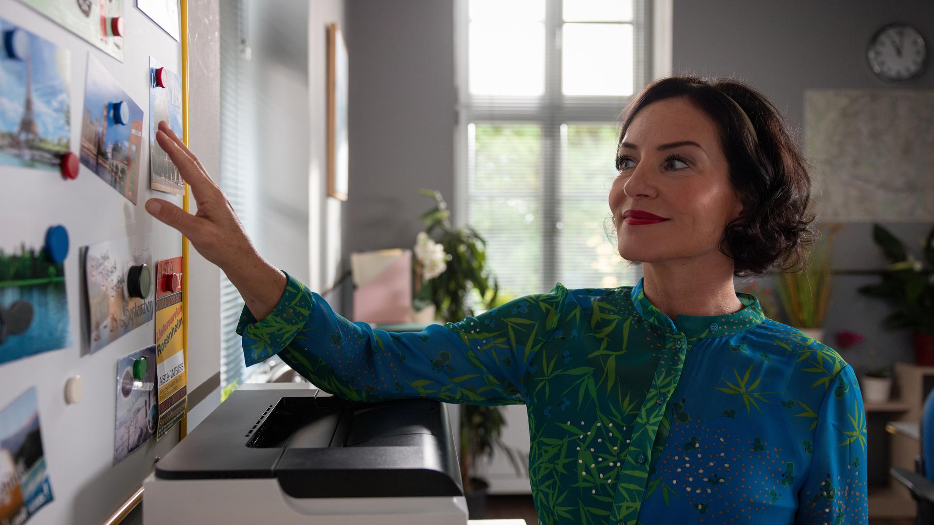 "Die Rosenheim-Cops - Mord und andere Ordnungswidrigkeiten": Miriam Stockl (Marisa Burger) steht lächelnd an der Magnettafel ihres Büros und hält ihre Hand an eine der angehefteten Postkarten. Unter der Tafel steht ein Drucker, daneben fällt Licht durch ein Fenster.