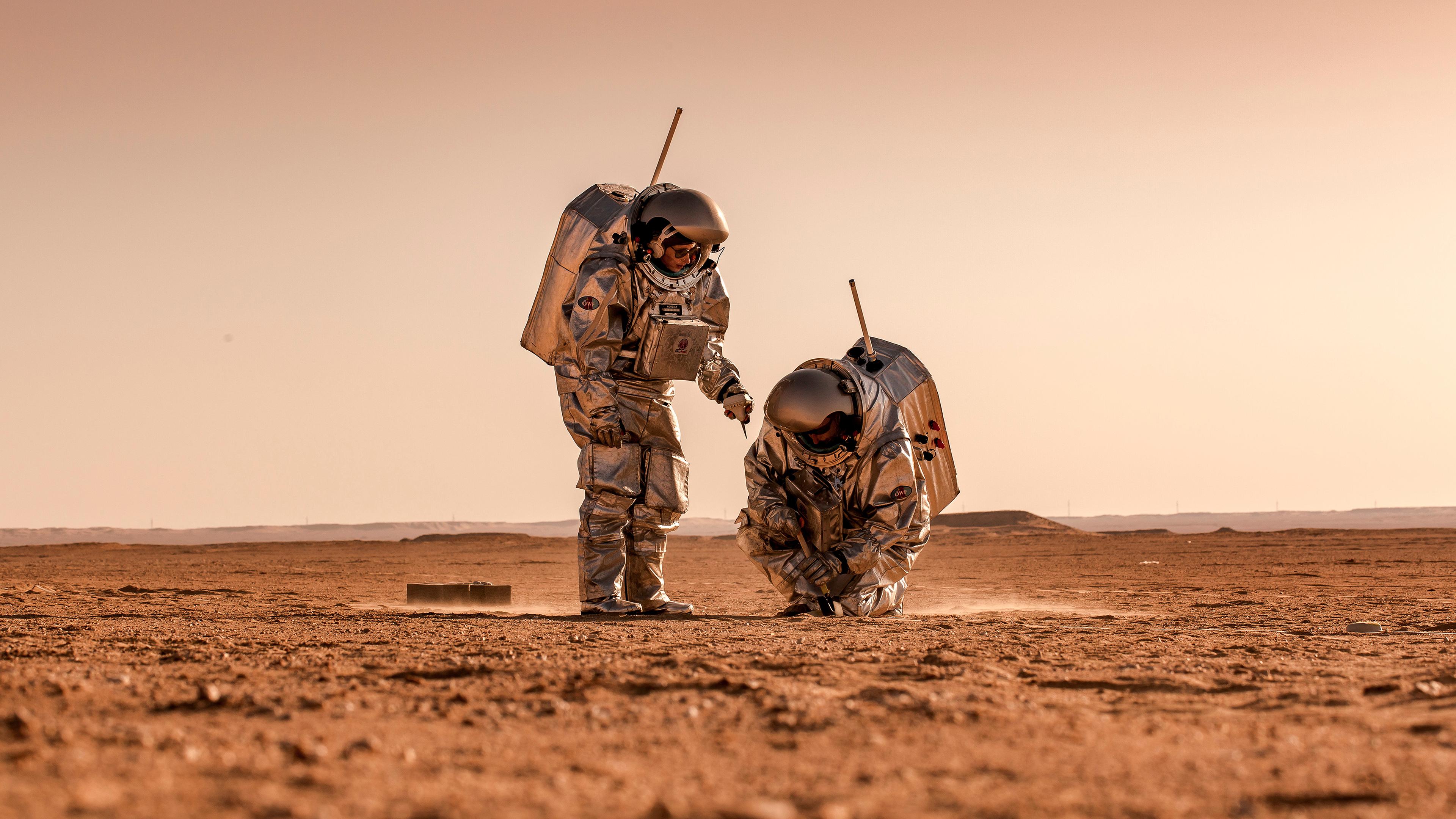 Zwei Personen in silbernen Raumanzügen nehmen Bodenproben in einer Wüste im Oman. Es handelt sich um eine Marssimulation.