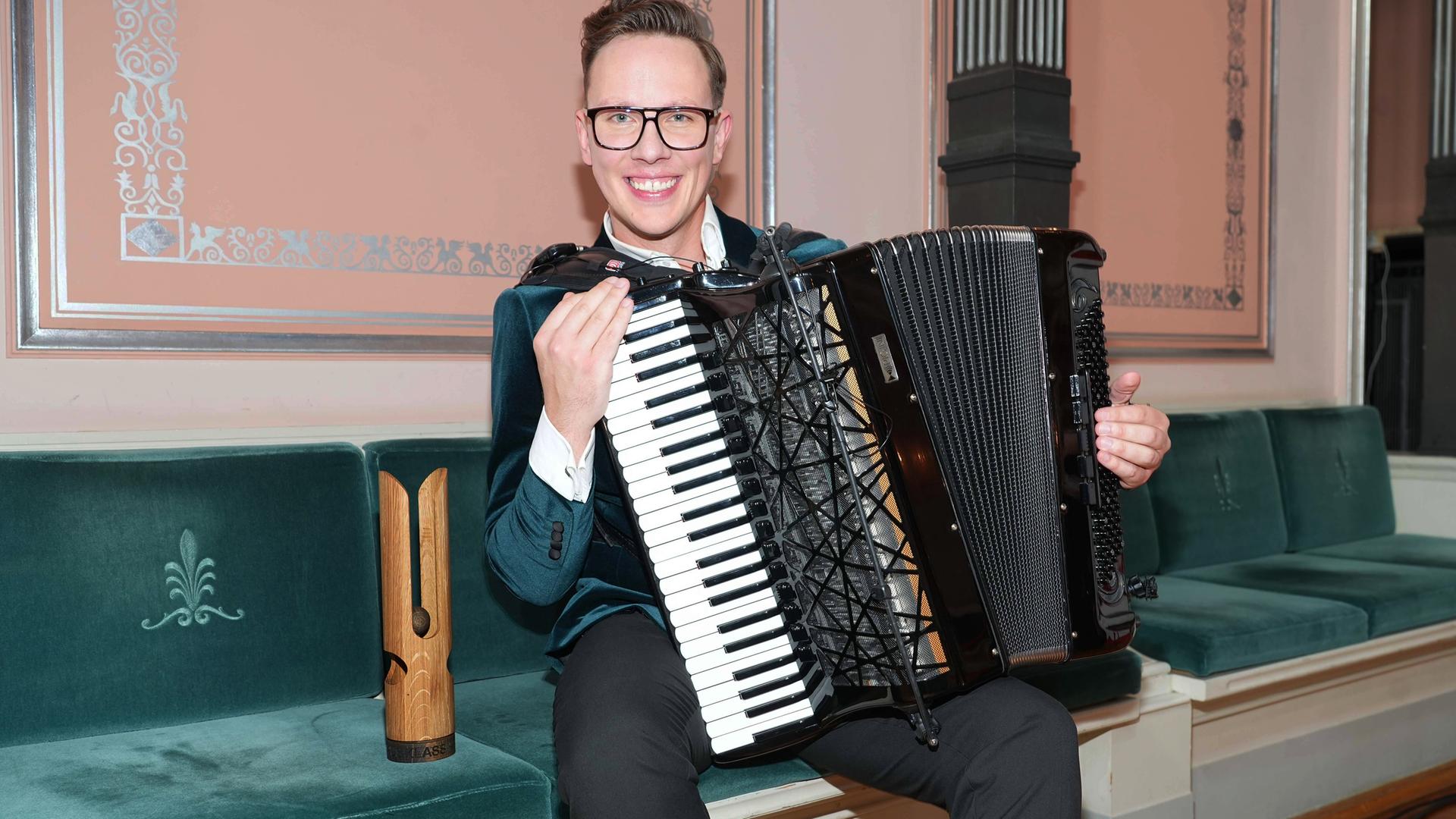 Ein junger Mann sitzt mit einem Akkordeon auf einer Bank. Neben ihm eine Trophäe aus Holz.