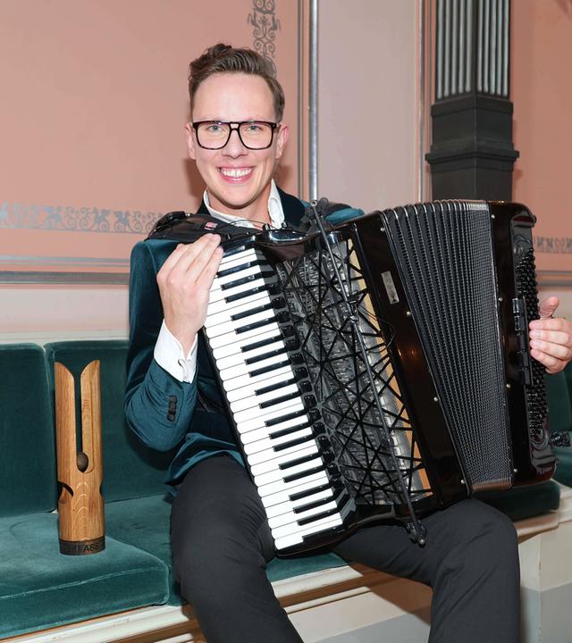 Ein junger Mann sitzt mit einem Akkordeon auf einer Bank. Neben ihm eine Trophäe aus Holz.