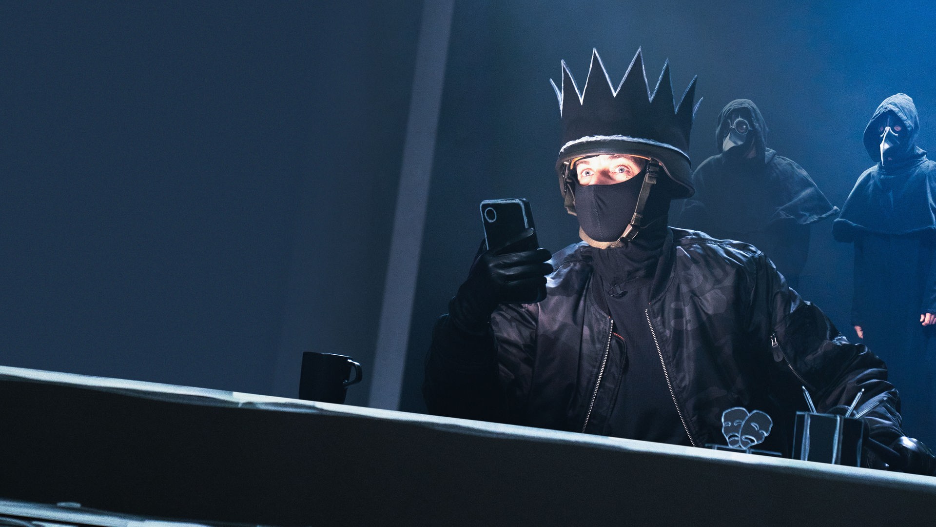 Auf einer Bühne sitzt im Vordergrund Jan Böhmermann, in schwarz gekleidet, mit einer schwarzen Krone und einer schwarzen Stoffmaske, die den unteren Teil seines Gesichts bedeckt. Er hält ein Smartphone in der Hand. Im Hintergrund stehen mehrere maskierte Personen in schwarzen Umhängen.