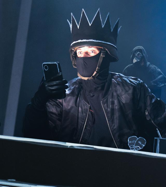 Auf einer Bühne sitzt im Vordergrund Jan Böhmermann, in schwarz gekleidet, mit einer schwarzen Krone und einer schwarzen Stoffmaske, die den unteren Teil seines Gesichts bedeckt. Er hält ein Smartphone in der Hand. Im Hintergrund stehen mehrere maskierte Personen in schwarzen Umhängen.