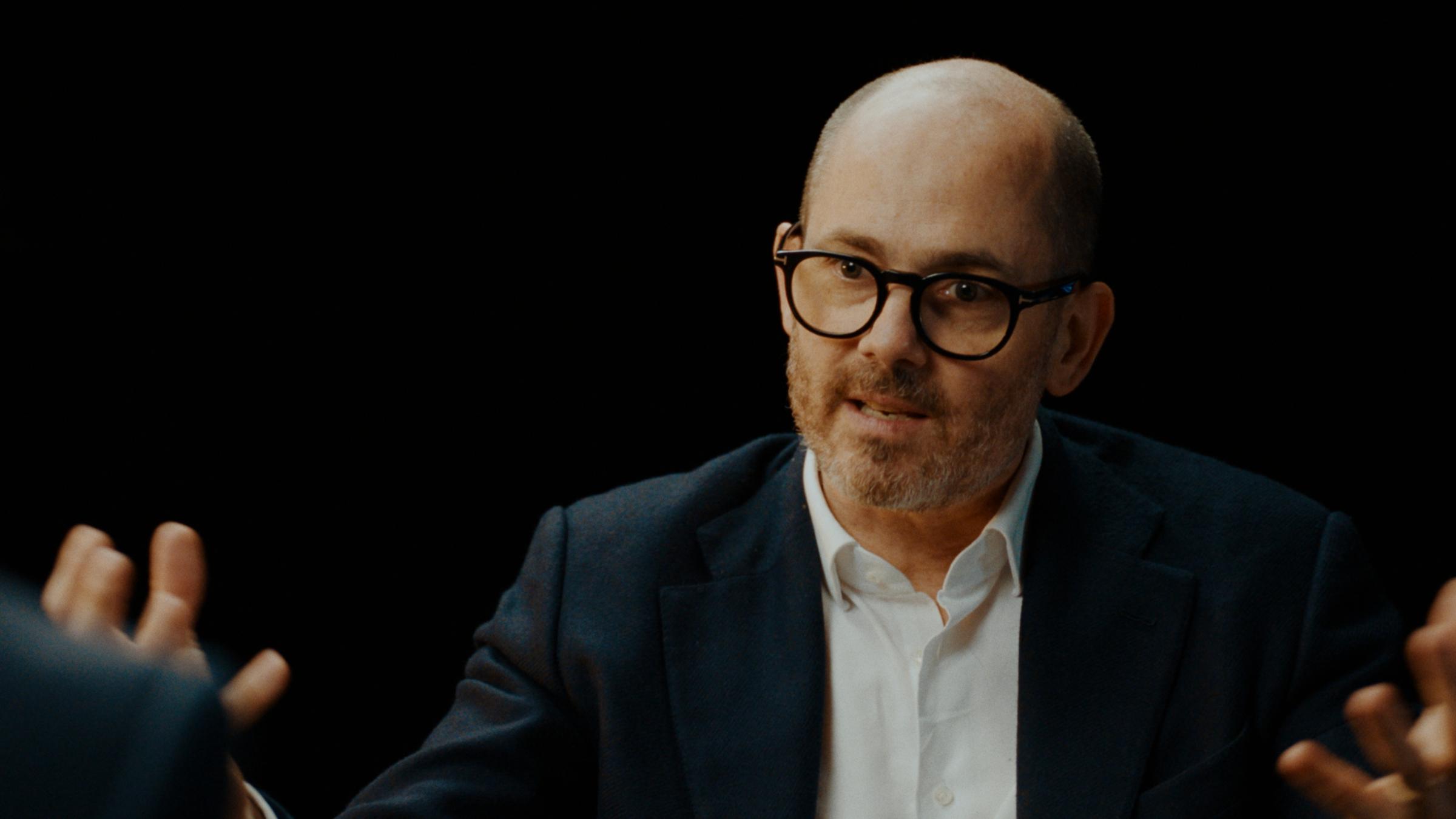 Mann in seinen frühen 50ern mit sehr kurzen grauen Haaren und gestutztem bronzenen Bart sitzt in einem braunen Sessel vor einem schwarzen Hintergrund. Er trägt eine schwarze Brille, ein dunkles Jackett und ein weißes Hemd.