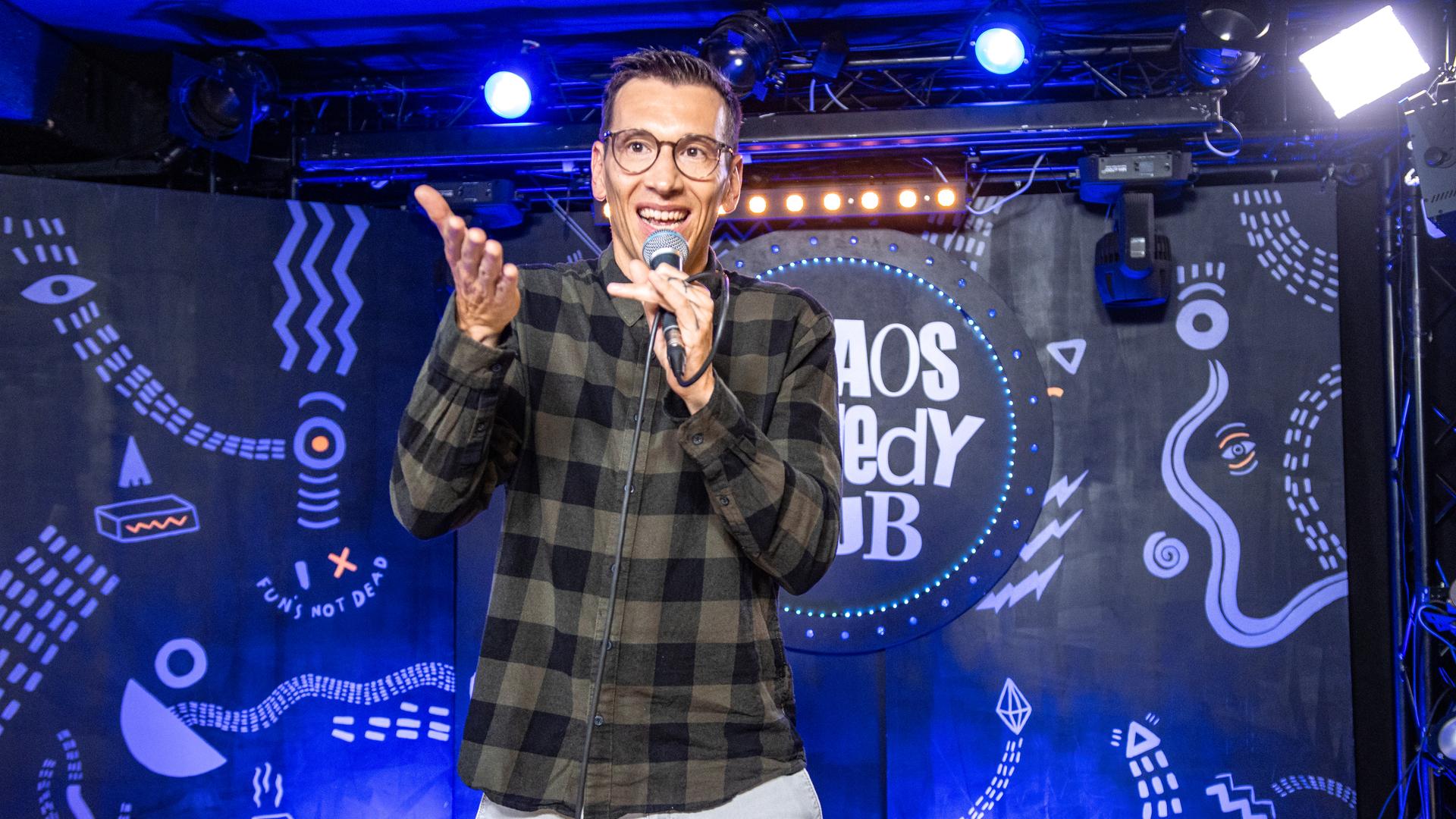 Mathias Albus steht in der Mitte des Bildes. In seiner linken Hand hält er ein Mikrofon. Er trägt ein kariertes Hemd.