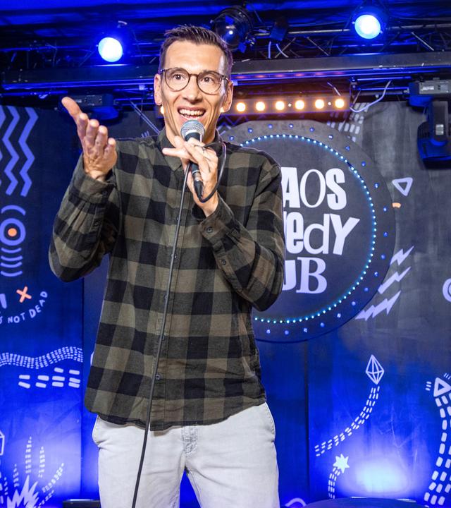 Mathias Albus steht in der Mitte des Bildes. In seiner linken Hand hält er ein Mikrofon. Er trägt ein kariertes Hemd.