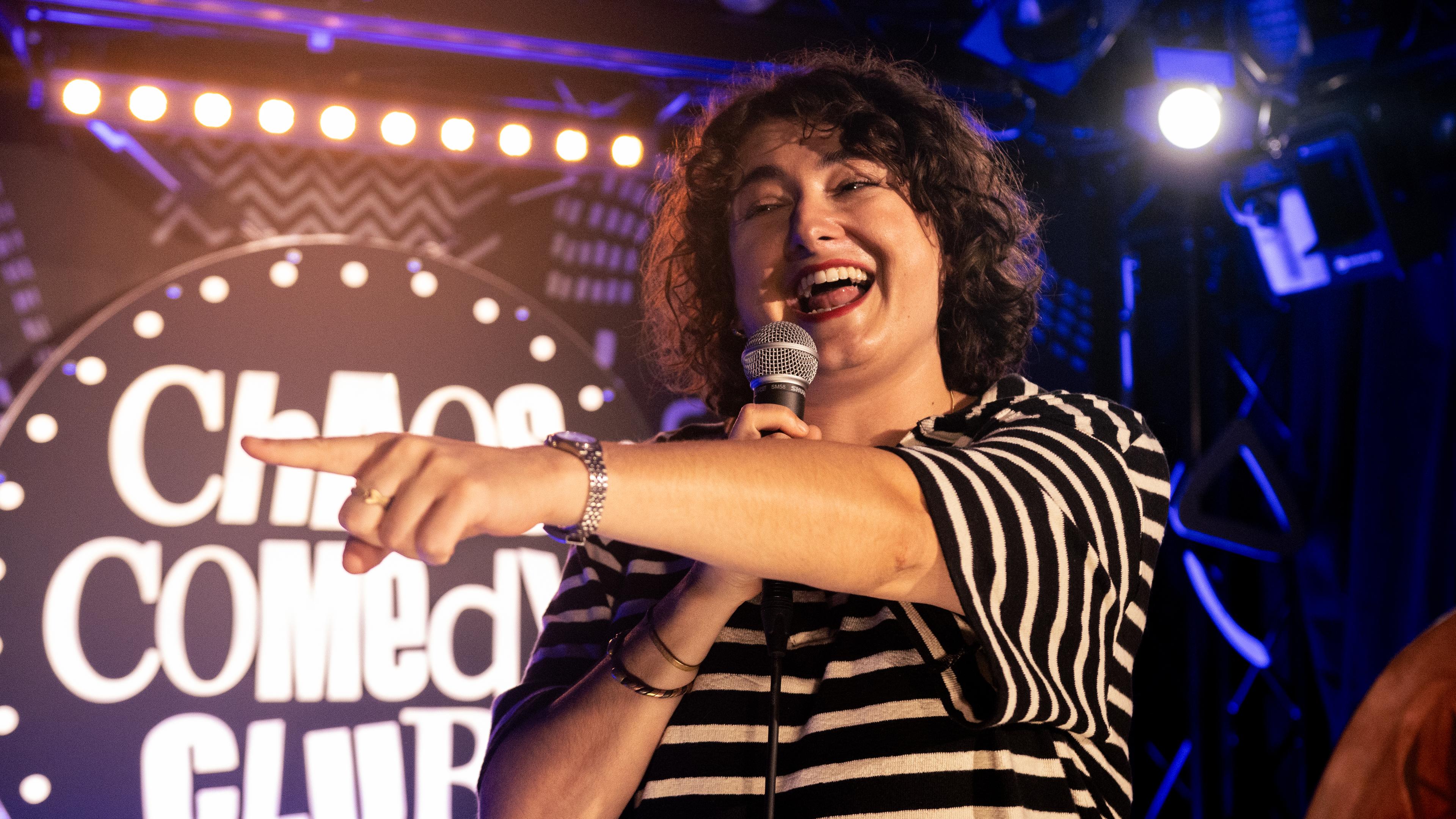 Matilde Keizer steht auf der Bühne. Sie deutet mit ihrem linken Arm in Richtung des linken Bildrandes. In der rechten Hand hält sie ein Mikrofon.