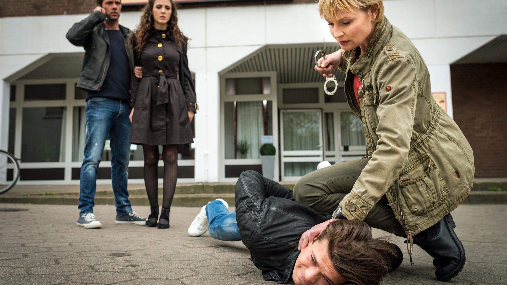 "Notruf Hafenkante - Matrjoschka": Boris Memedow (Eugen Bauder) liegt auf dem Boden. Melanie Hansen (Sanna Englund) sitzt in der Hocke neben ihm und hat ihr Knie zur Fixierung auf seinem Rücken. Mit der linken Hand drückt sie seinen Kopf auf den Boden in der Rechten hält sie Handschellen, die sie im Begriff ist ihm anzulegen. Im Hintergrund stehen Mattes Seeler (Matthias Schloo) und Marina Petrowa (Katharina Rivilis) nebeneinander. Mattes hält Marina an ihrem rechten Arm. Er telefoniert mit dem Handy. Mattes und Melanie sind beide in Zivilkleindung.