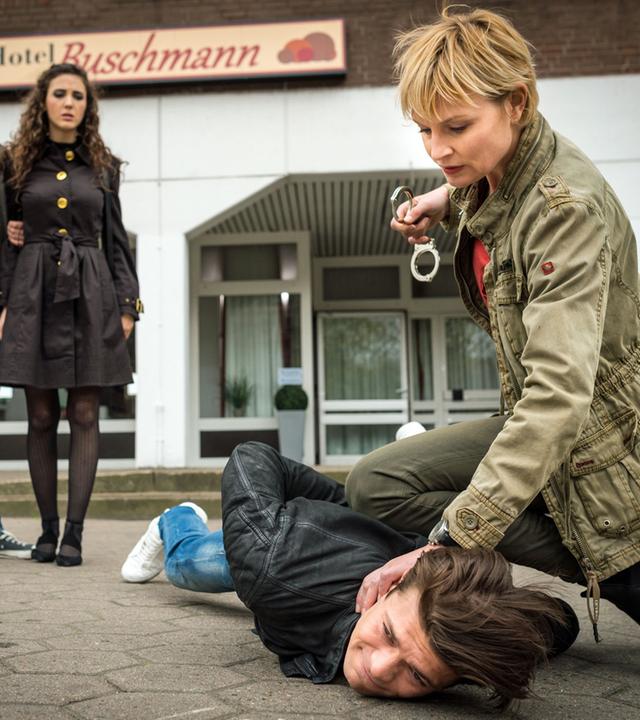 "Notruf Hafenkante - Matrjoschka": Boris Memedow (Eugen Bauder) liegt auf dem Boden. Melanie Hansen (Sanna Englund) sitzt in der Hocke neben ihm und hat ihr Knie zur Fixierung auf seinem Rücken. Mit der linken Hand drückt sie seinen Kopf auf den Boden in der Rechten hält sie Handschellen, die sie im Begriff ist ihm anzulegen. Im Hintergrund stehen Mattes Seeler (Matthias Schloo) und Marina Petrowa (Katharina Rivilis) nebeneinander. Mattes hält Marina an ihrem rechten Arm. Er telefoniert mit dem Handy. Mattes und Melanie sind beide in Zivilkleindung.