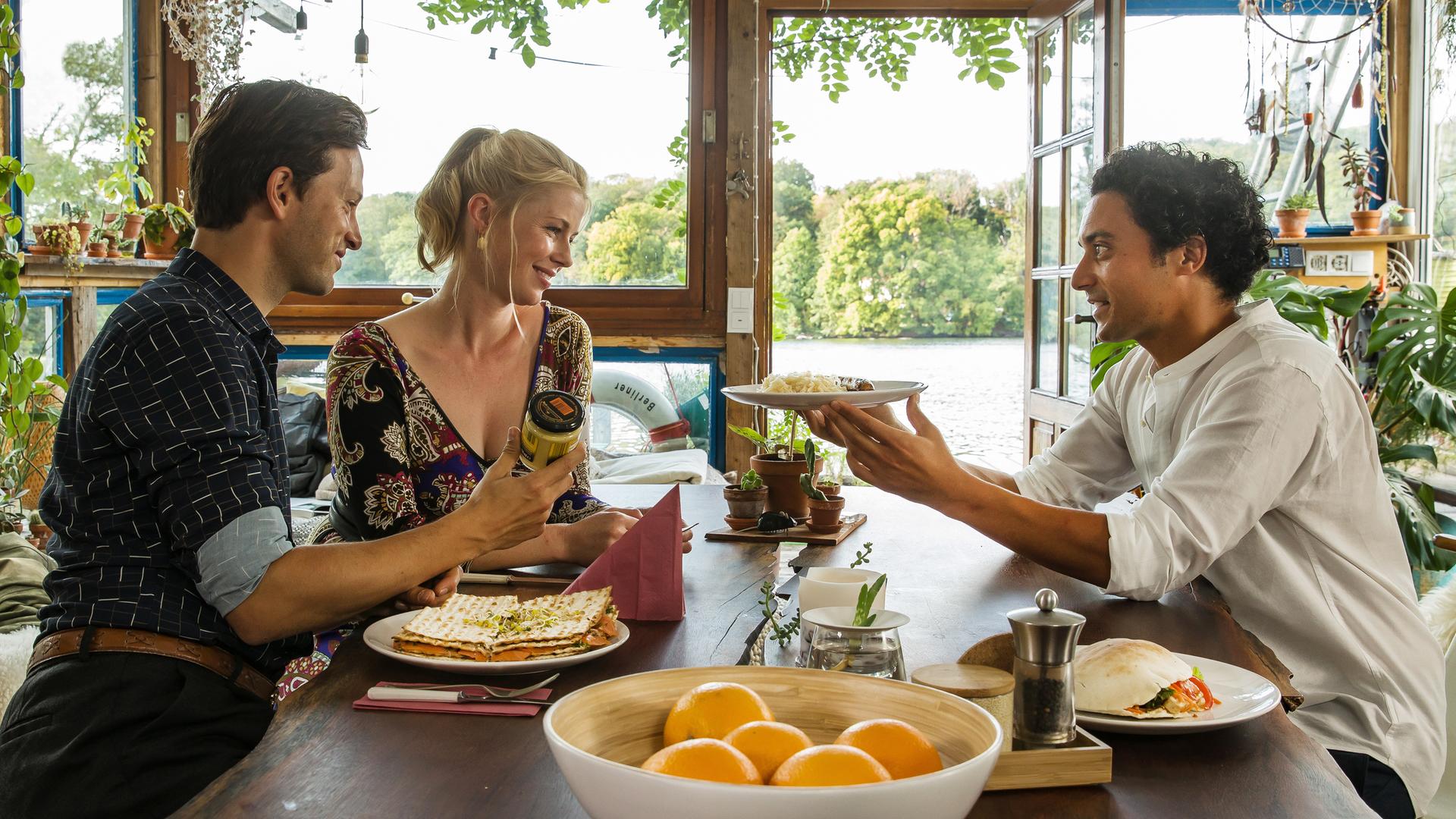 "Matze, Kebab und Sauerkraut": Noah (Franz Dinda), Charlottes (Christine Eixenberger) und Hakim (Omar El-Saeidi) sitzen gemeinsam am Tisch. Hakim hält Charlotte einen Teller hin, im Hintergrund glitzert der See